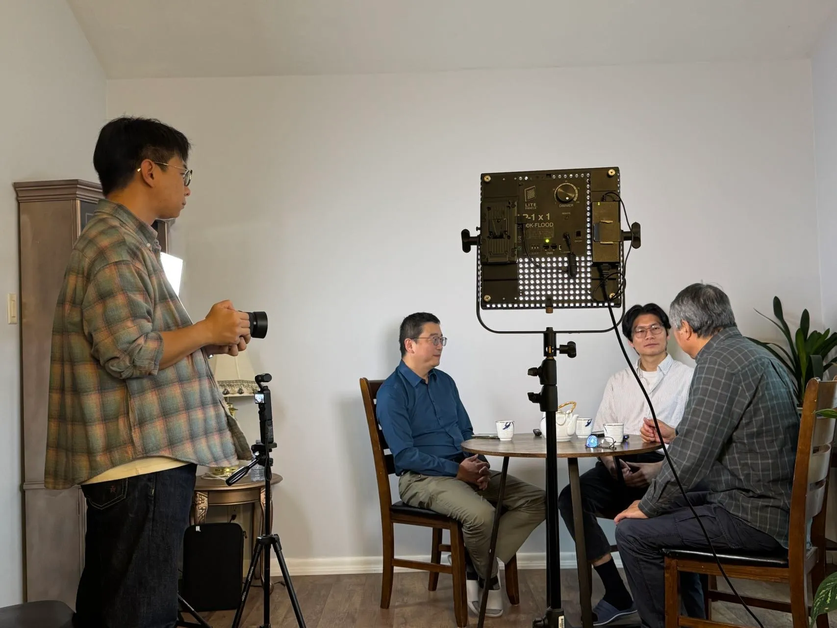Ning operating a camera while the creator interviews two guests at a table, lit by a panel light