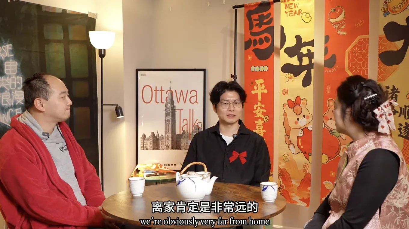 A still from an Ottawa Talks episode: three people talk around a small table with a teapot, and the framed poster hangs on the wall behind them