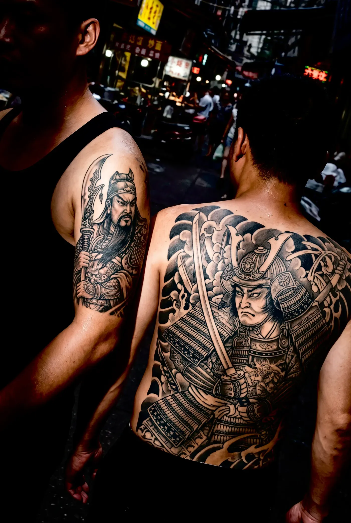 AI-generated street photo of two men at a night market, one shirtless, whose tattoos of armoured warriors on back and arm seem to face each other
