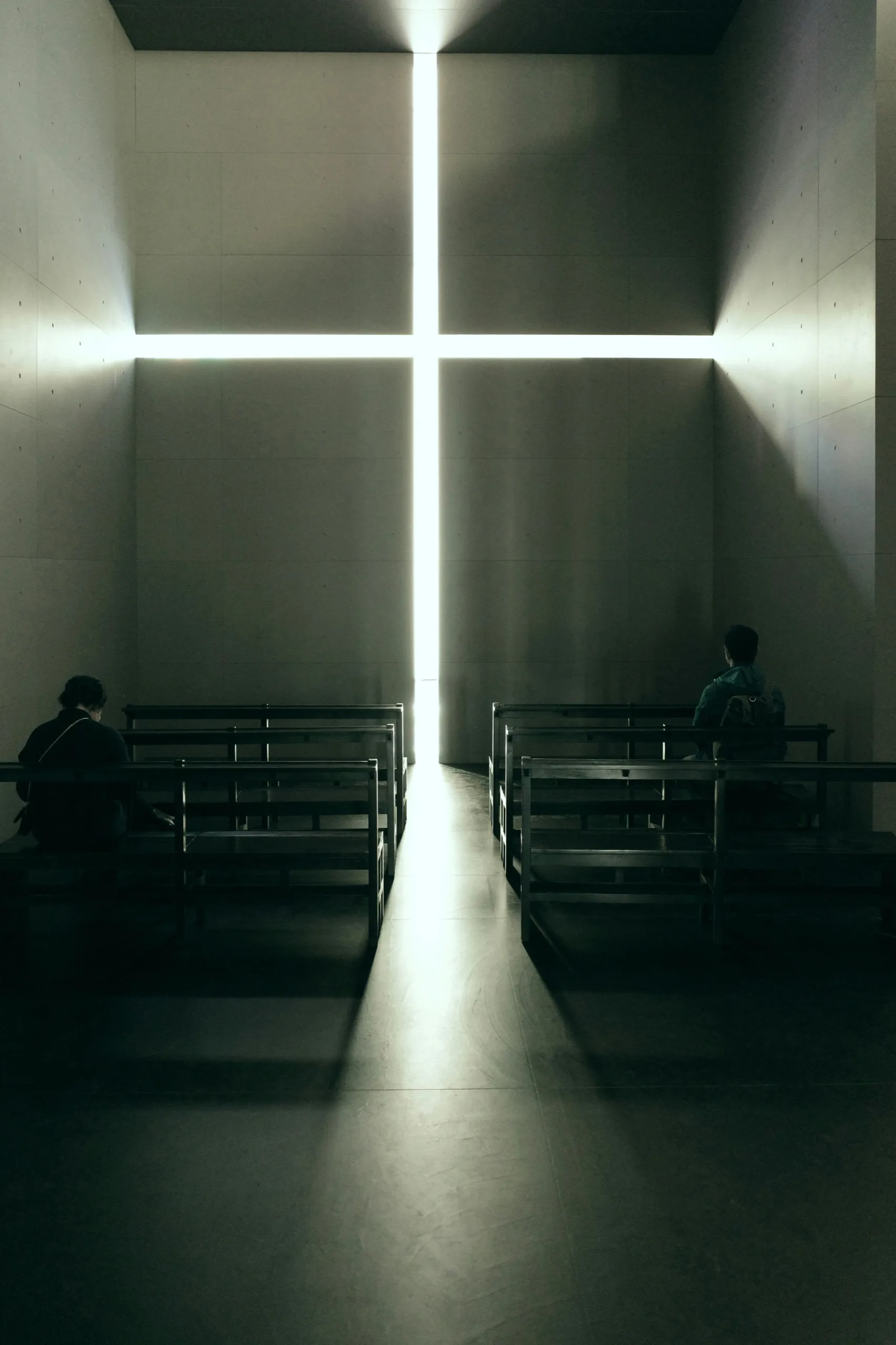 Two people seated on pews below a cross of light in a concrete chapel
