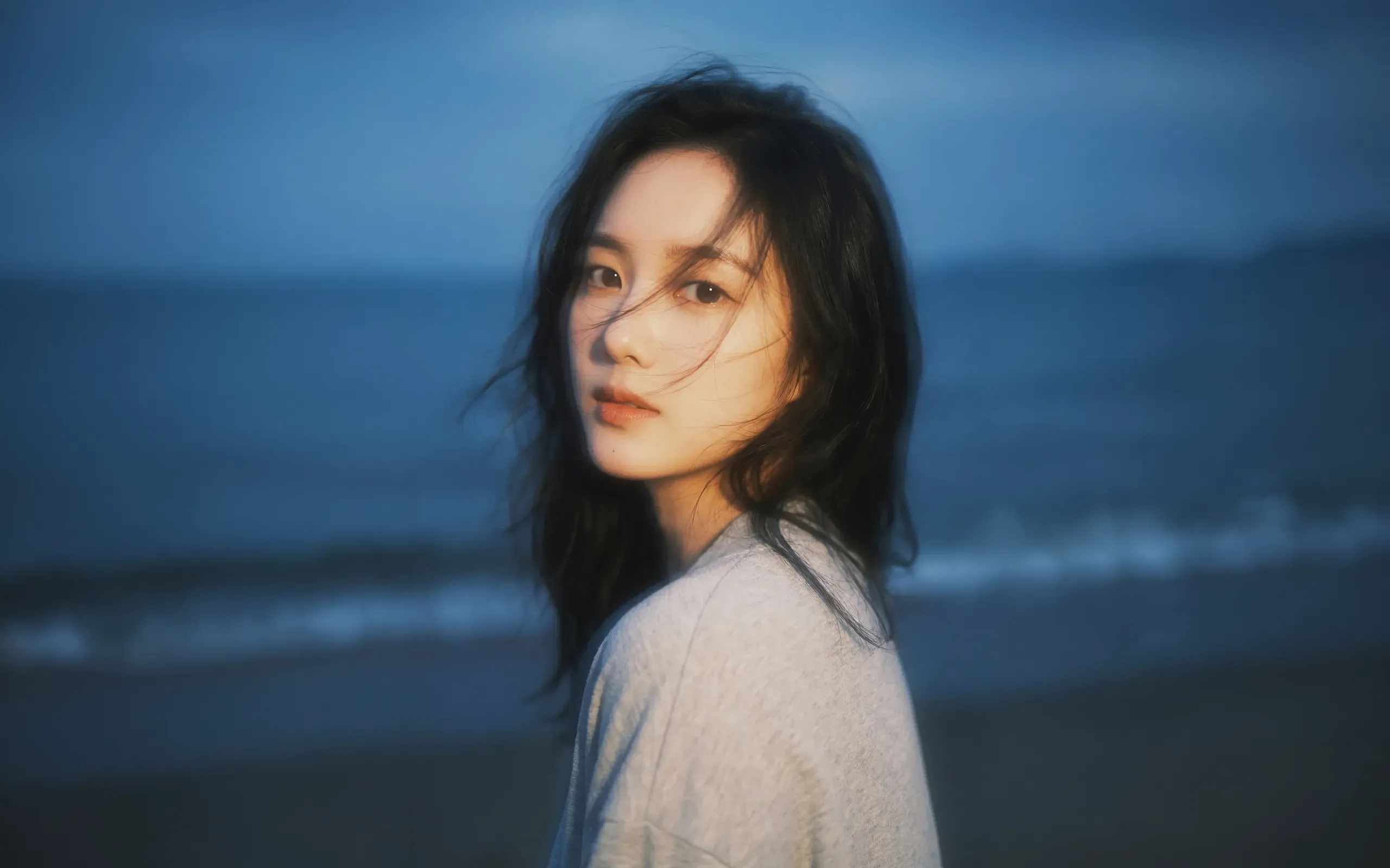 AI-generated portrait of a woman in a pale shirt on a beach at dusk, glancing back as the wind lifts her hair