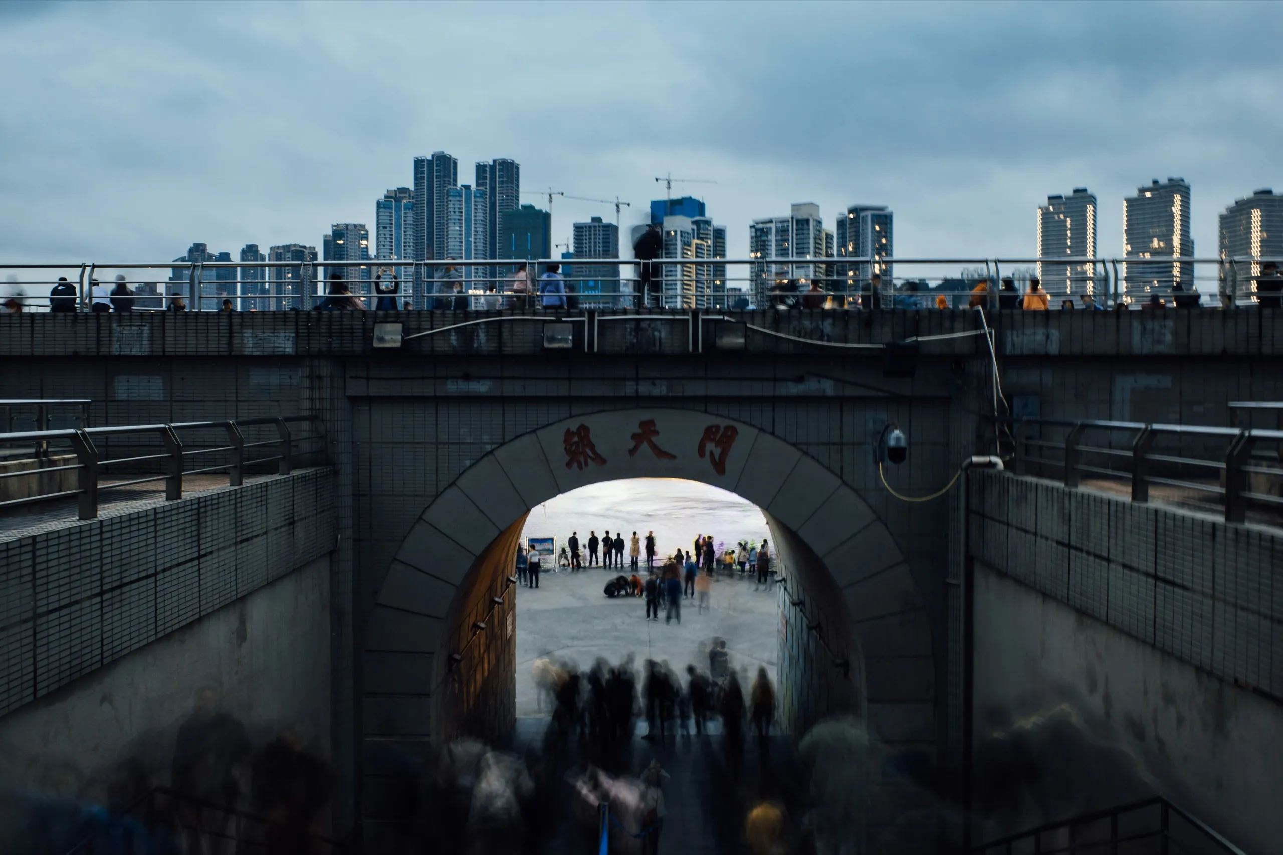A crowd blurred in motion walking through an arch beneath a bridge, towers behind