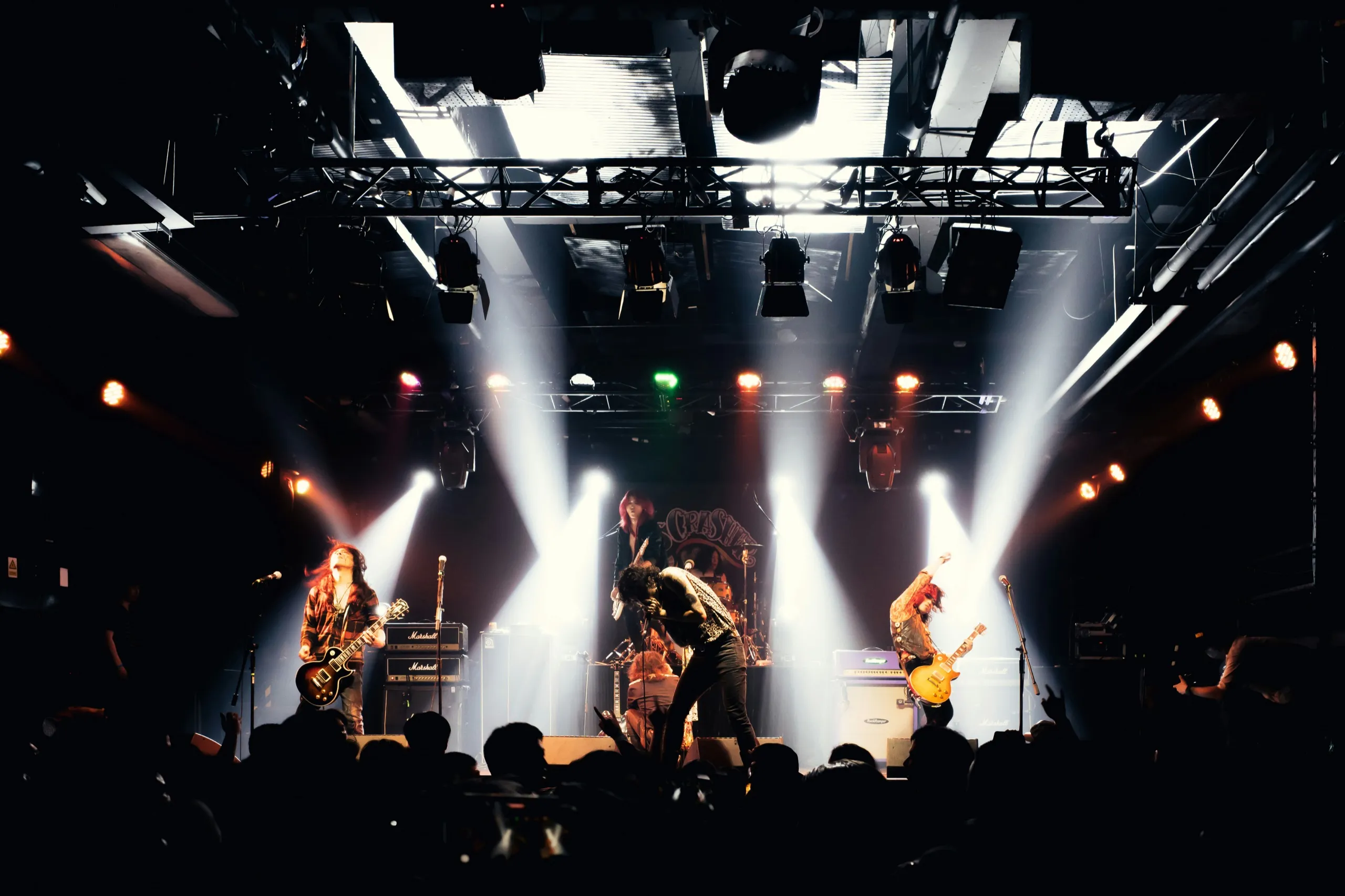 A rock band under white spotlight beams, seen over the crowd