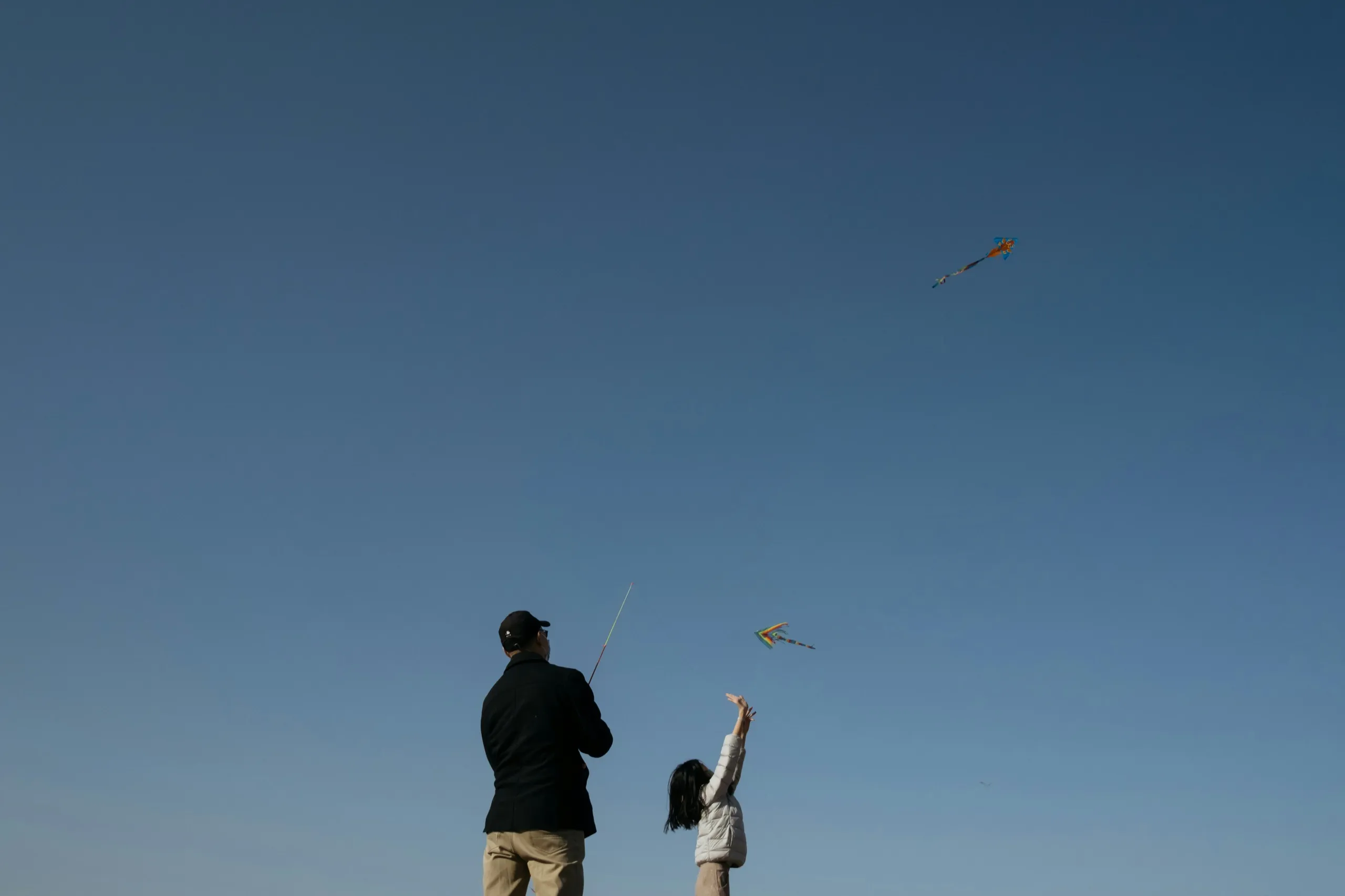 A man and a little girl flying kites against a clear blue sky