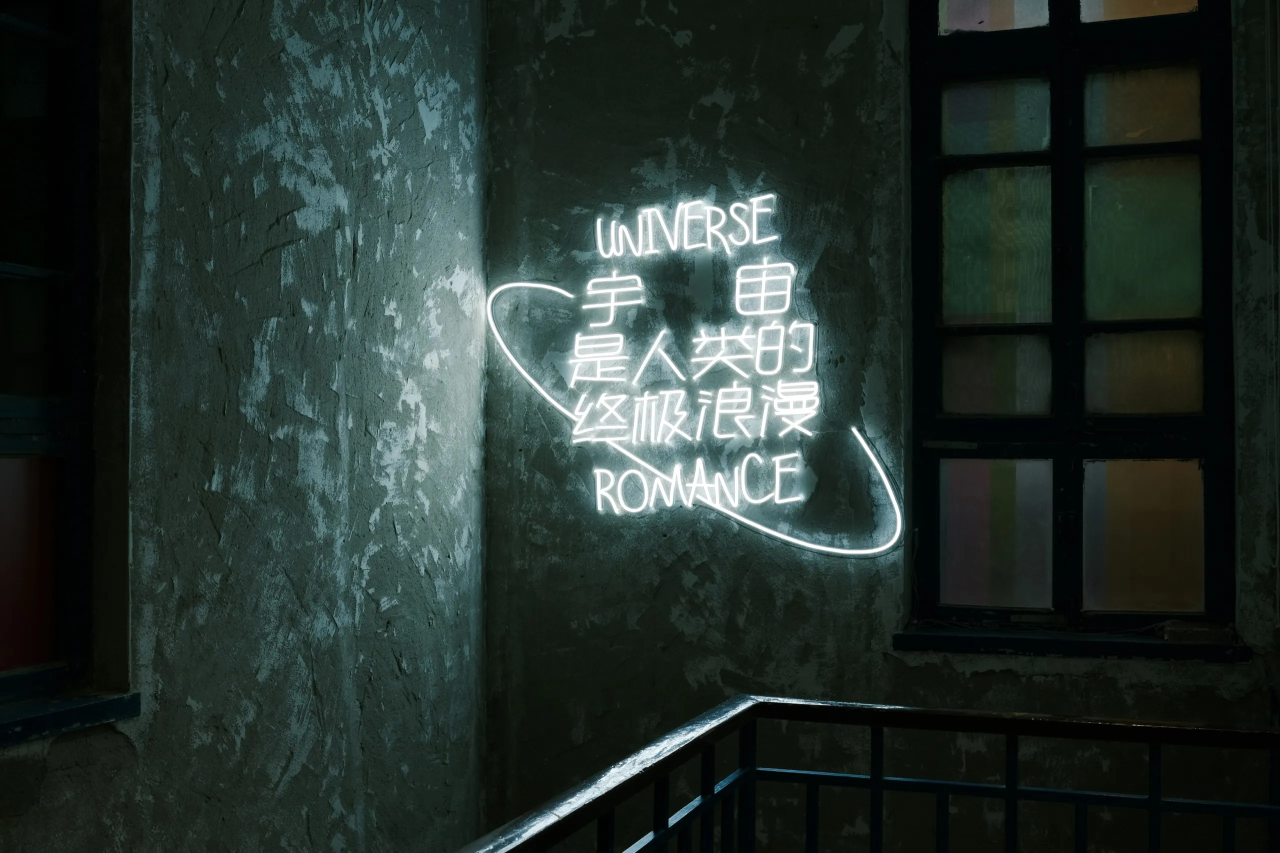 A white neon sign reading Universe is the ultimate romance of humankind, in English and Chinese, on a weathered stairwell wall