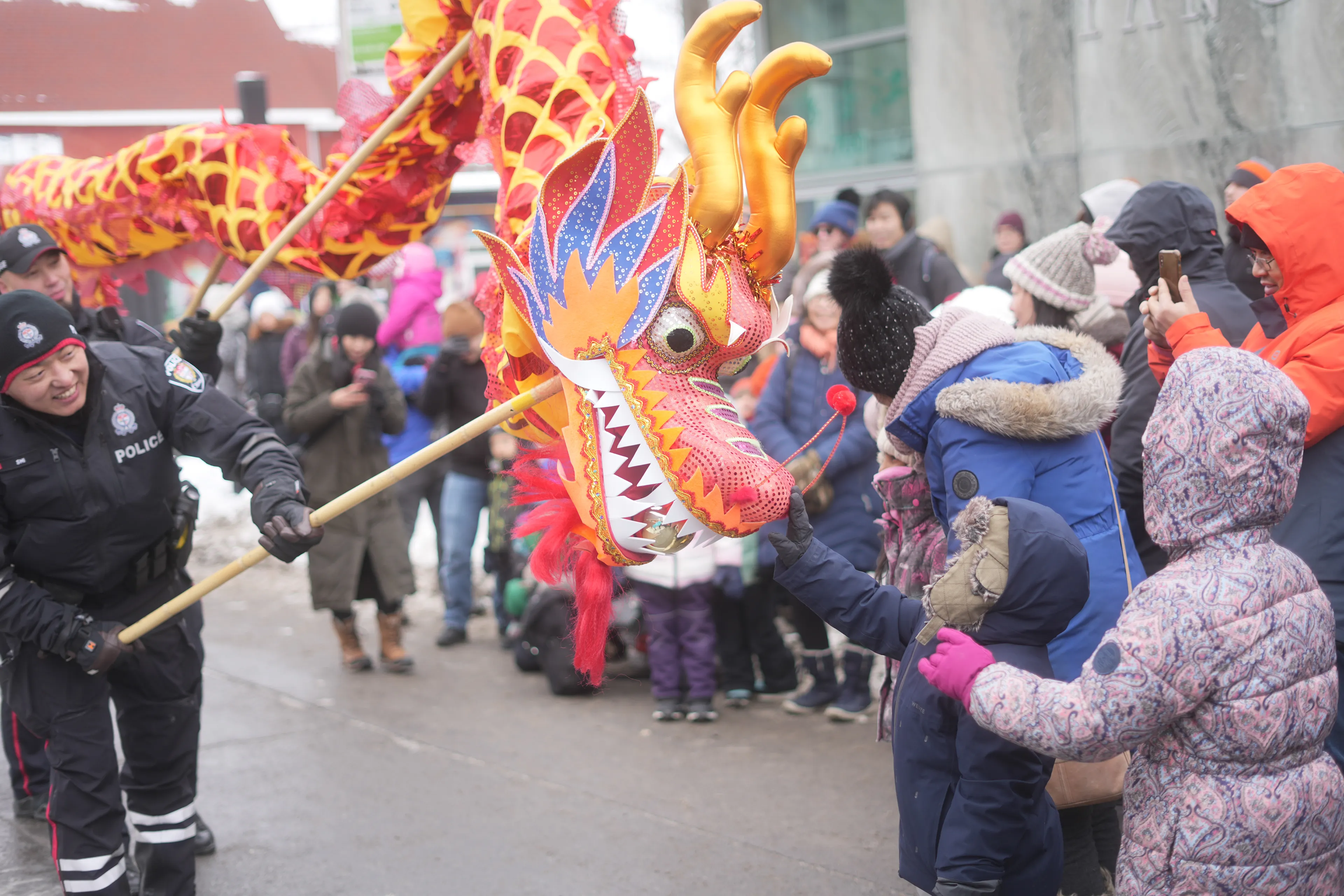 A red and gold dragon leans toward children in winter coats at the Ottawa Chinatown New Year Parade, with a police officer beside it.